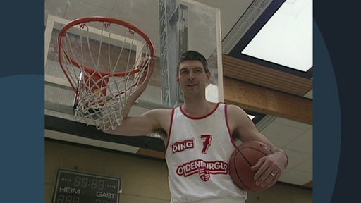Der ehemalige Baskets Oldenburg Spieler Mark Smed hält mit seiner Hand einen Basketballkorb und seine andere Hand hält einen Basketball. 
