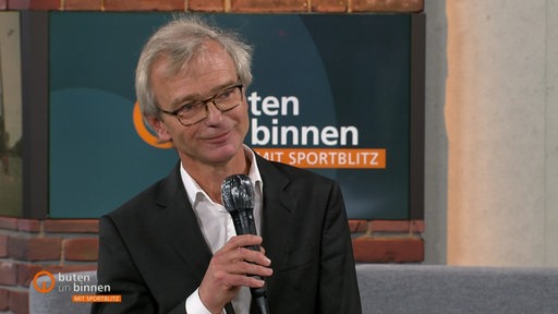 Ein Mann mit Brille und grauen Haaren hält in einem TV-Studio ein Mikrofon in der Hand
