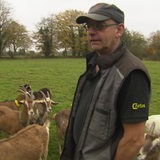 Ein Mann mit Cappy und Brille schaut in die Kamera - hinter ihm steht eine Ziegenherde