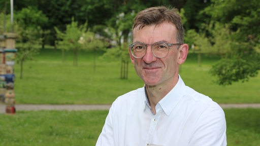Mann mit Brille in weißem Hemd vor Parklandschaft lächelt für Portrait in Kamera