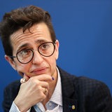 Eine Frau mit kurzen Haaren und großer Brille stützt ihren Kopf auf ihre Hand.