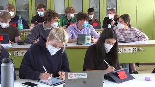 Schüler und Schülerinnen einer Oberstufenklasse sitzen zusammen im Unterricht und tragen medizinische Masken.