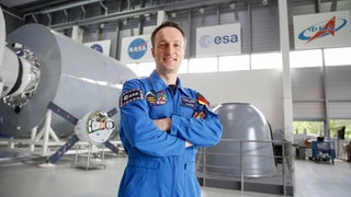 Der Astronaut Matthias Maurer steht in einem blauen Overall in einer großen Halle