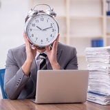 Ein Mann in einem Büro hält sich einen Wecker vors Gesicht (Symbolbild)