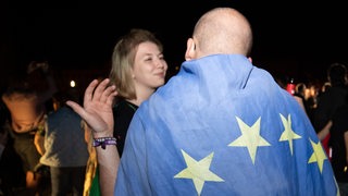 Ein Mann trägt eine Europaflagge über den Schultern und unterhält sich mit einer lächelnden Frau.