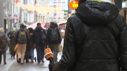Eine Frau mit dunkler Winterjacke von hinten hält ein Mikrofon in der Hand, im Hintergrund zahlreiche flanierende Menschen.