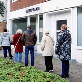 Patienten stehen vor einer Arztpraxis Schlange.