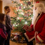 Der Weihnachtsmann sitzt zur Bescherung in einem Haus neben einem geschmückten Weihnachtsbaum während zwei Mädchen ihm ein Lied vorsingen