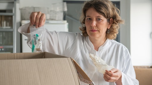 Eine Frau im Laborkittel hält Tüten mit Plastikteilen in den Händen.