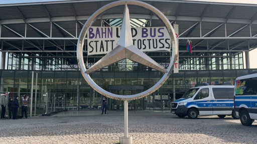Der große Stern vor einem Mercedes Benz Werk, im Hintergrund ist ein großes Protestplakat gegen Autos aufgehängt.