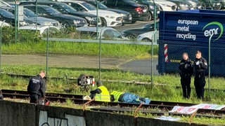 Menschen sitzen mit Warnwesten auf Gleisen. Um sie herum stehen drei Polizisten