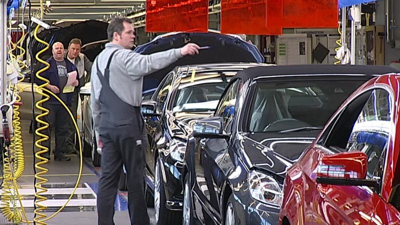 Drei Mercedes Mitarbeiter stehen an einem Fließband, auf dem mehrere Autos stehen.