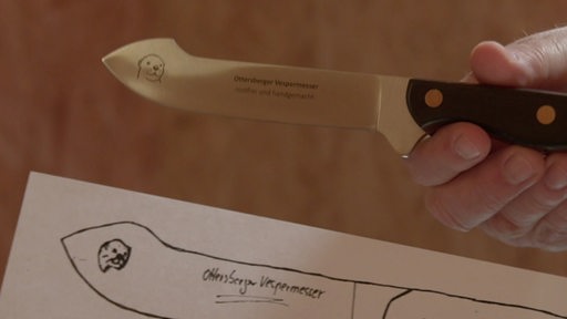 Eine Hand hält ein Messer auf dem ein Schriftzug und ein Otter eingraviert sind. Darunter sieht man das Blatt Papier mit der Skizze für die Sonderanfertigung.