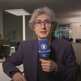 Ein Mann mit einem blauen Mikrofon sitzt an einem Schreibtisch neben einem Bildschirm mit einer Wetterkarte.