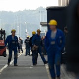 Arbeiter in blauen Overalls und mit gelben Helmen gehen über ein Betriebsgelände.
