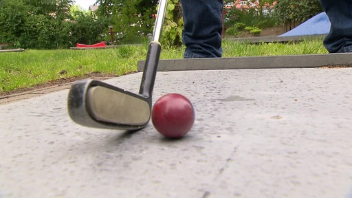 Eine Nahaufnahme eines Minigolf-Schlägers, der auf dem Boden an einen Minigolf-Ball gehalten wird.