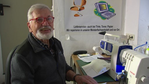 Ein älterer Mann mit weißen Haaren und roter Brille lächelt, vor ihm steht auf dem Tisch eine Nähmaschine