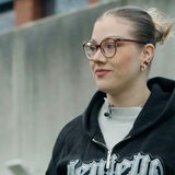 Eine junge Frau steht vor einem grauen Gebäude und schaut schräg nach links.