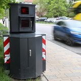 Ein mobiler Blitzer-Anhänger steht an einer Straße, ein Auto und Bus fahren nebenher