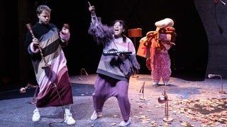 Theaterszene mit drei Akteuren in lilafarbenem Bühnenlich