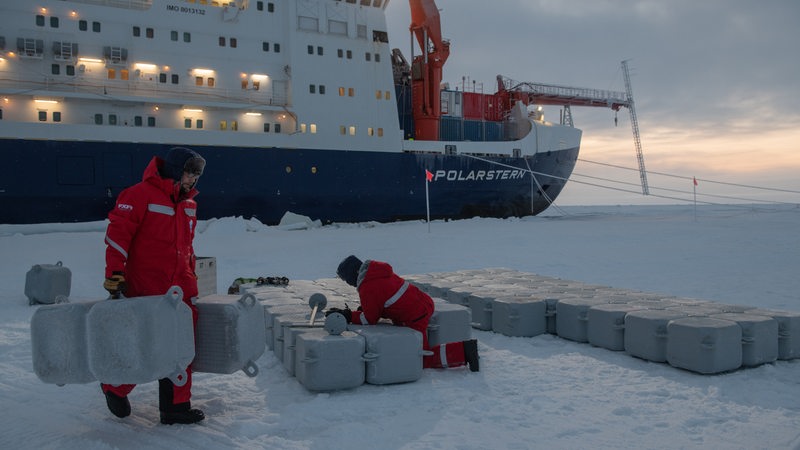 Zwei Personen tragen Ausrüstung aufs Eis. Im Hintergrund liegt die Polarstern.