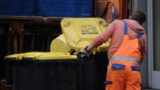 Ein Mitarbeiter der Müllabfuhr schiebt mehrere Gelbe Tonnen zu einem Müllwagen.