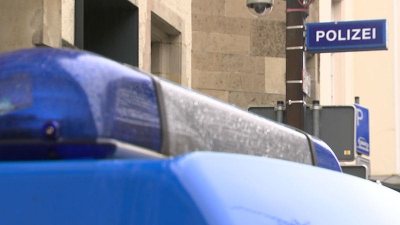 Ein Polizeiauto mit Blaulicht auf dem Dach steht vor einen Polizeiwache. Im Hintergrund ist ein Polizeischild montiert. 