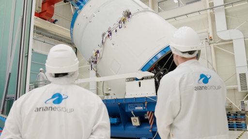 Zwei Arbeiter gucken sich die Verladung der Ariane 5 Rakete an. 