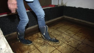 Jemand tritt mit Gummistiefeln in eine Wasserfläche in einem Keller.
