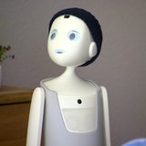 Ein Roboter ist von der Seite zu sehen, im Vordergrund unscharf ein Arm