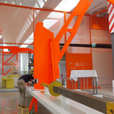Ein Mann steht auf einem Hocker und schraubt an einem orangefarbenen Ausstellungsstück.
