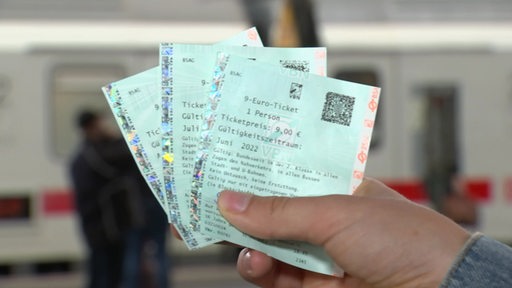 Eine Hand hällt drei Neun-Euro-Tickets vor einem Zug.