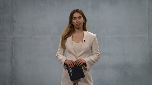 Eine junge Frau steht mit Moderationskarten in der Hand vor einer grauen Wand.