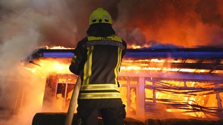 Ein Feuerwehrmann von hinten betrachtet hält einen Wasserschlauch und richtet ihn auf eine Gebäude, das in Flammen steht.