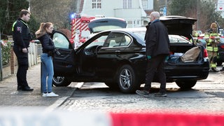 Ein schwarzes Auto steht an einem Straßenrand. Zwei Männer und eine Frau stehen davor. Im Hintergrund ist ein Feuerwehrmann zu sehen.