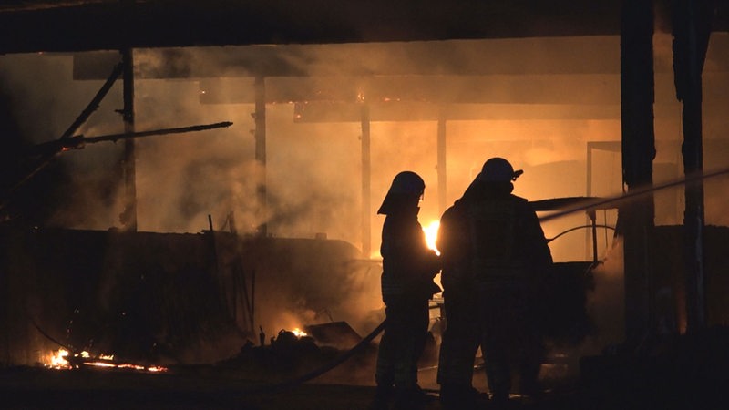 Zwei Feuerwehrleute stehen vor einem brennenden Gebäude.