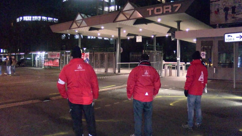 Drei Männer in Jacken von IG Metall stehen vor dem Tor 7 des Bremer Mercedes Benz-Werks.
