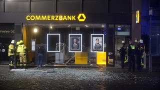 Feuerwehrleute stehen vor einer Filiale der Commerzbank, deren Glasfassade geborsten ist.