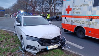 Im Vordergrund ein zerstörtes Auto. Im Hintergrund Polizisten, ein Rettungswagen und ein weiterer Unfallwagen.