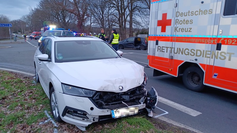 Im Vordergrund ein zerstörtes Auto. Im Hintergrund Polizisten, ein Rettungswagen und ein weiterer Unfallwagen.