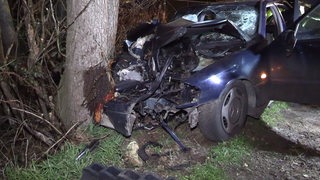 Ein komplett zerstörtes Auto ist gegen einen Baum gedrückt.
