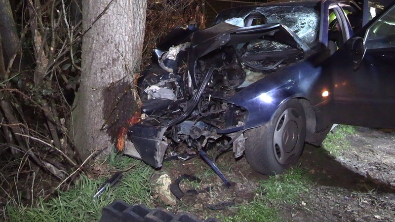 Ein komplett zerstörtes Auto ist gegen einen Baum gedrückt.