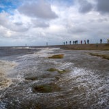 Wellen schlagen an den Nordseestrand, im Hintergrund stehen Menschen auf einem Damm.