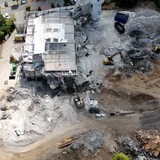 Ein Gebäude wird abgerissen. Auf der Baustelle sind um das Gebäude mehrere Bagger zu sehen.