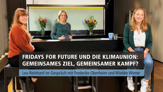Drei Frauen sitzen an einem schwarzen Tresen und schauen in die Kamera, die ihnen frontal gegenübersteht.