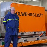 Ein Mann in blauer Uniform steht vor einer orangen Plane mit der Aufschrift "Ölwehrgerät".