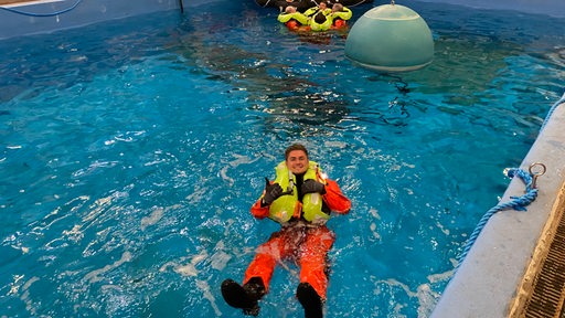 Ein Mann in orangefarbenem Sicherheitsanzug und gelber Sicherheitsweste treibt in einem Schwimmbecken.