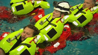 Vier Personen in roten Schutzanzügen und mit gelben Rettungswesten treiben in einem Schwimmbecken.