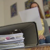 Im Vordergrund ist ein Ordner und im Hintergrund eine Frau, welche auf ein Blattpapier guckt, zu sehen.