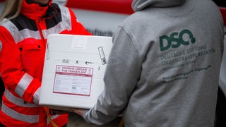 Eine Mitarbeiterin des Deutschen Roten Kreuzes nimmt eine Transportbox für ein menschliches Organ für eine Transplantation von einem Koordinator der Deutschen Stiftung Organtransplantation entgegen.
